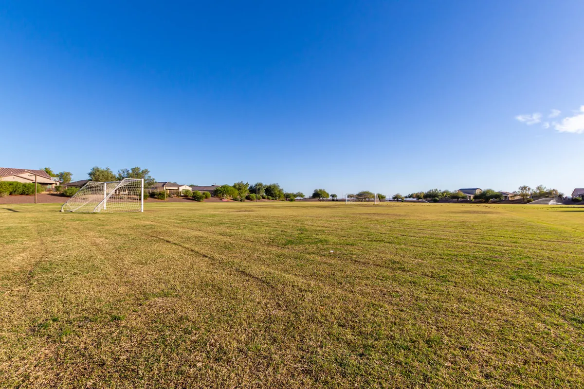 A vibrant outdoor soccer field surrounded by lush grass and nearby homes, with soccer goals set against a clear blue sky. The area is alive with community energy, suggesting youth sports leagues, informal matches, and gatherings.