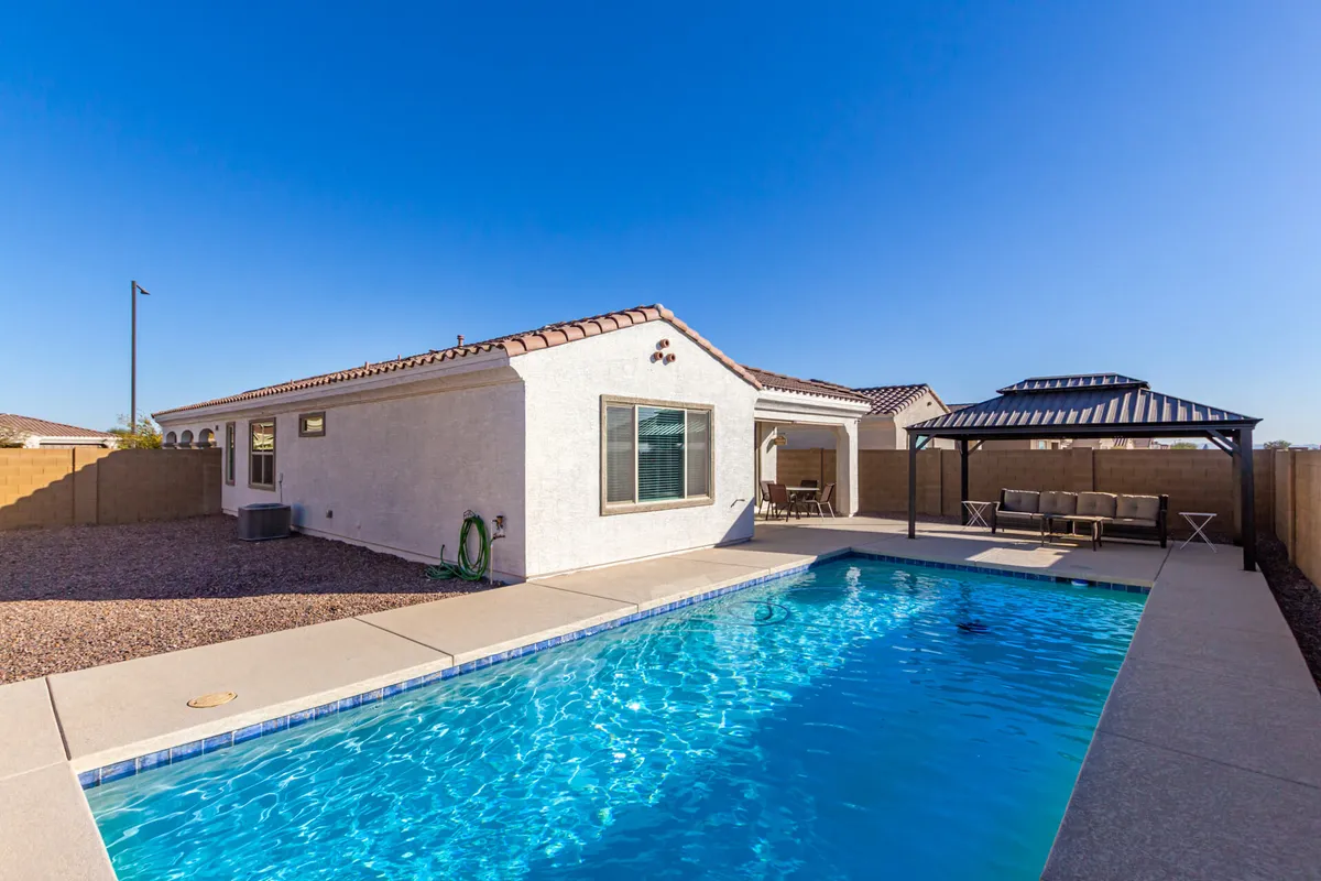 This exterior image captures a sparkling swimming pool bordered by a covered patio and the house's exterior. The patio is set up for outdoor relaxation, bathed in sunlight, and the clear blue sky reinforces the sense of tranquility and desert sunshine.