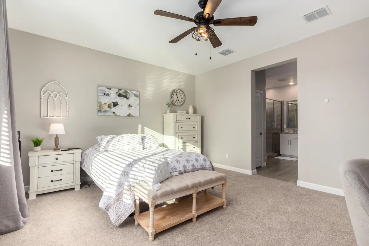 A serene bedroom featuring a comfortable bed, ceiling fan, nightstand with a lamp, Southwestern-inspired artwork, and a bench at the foot of the bed. The room is softly lit, inviting relaxation and rest.