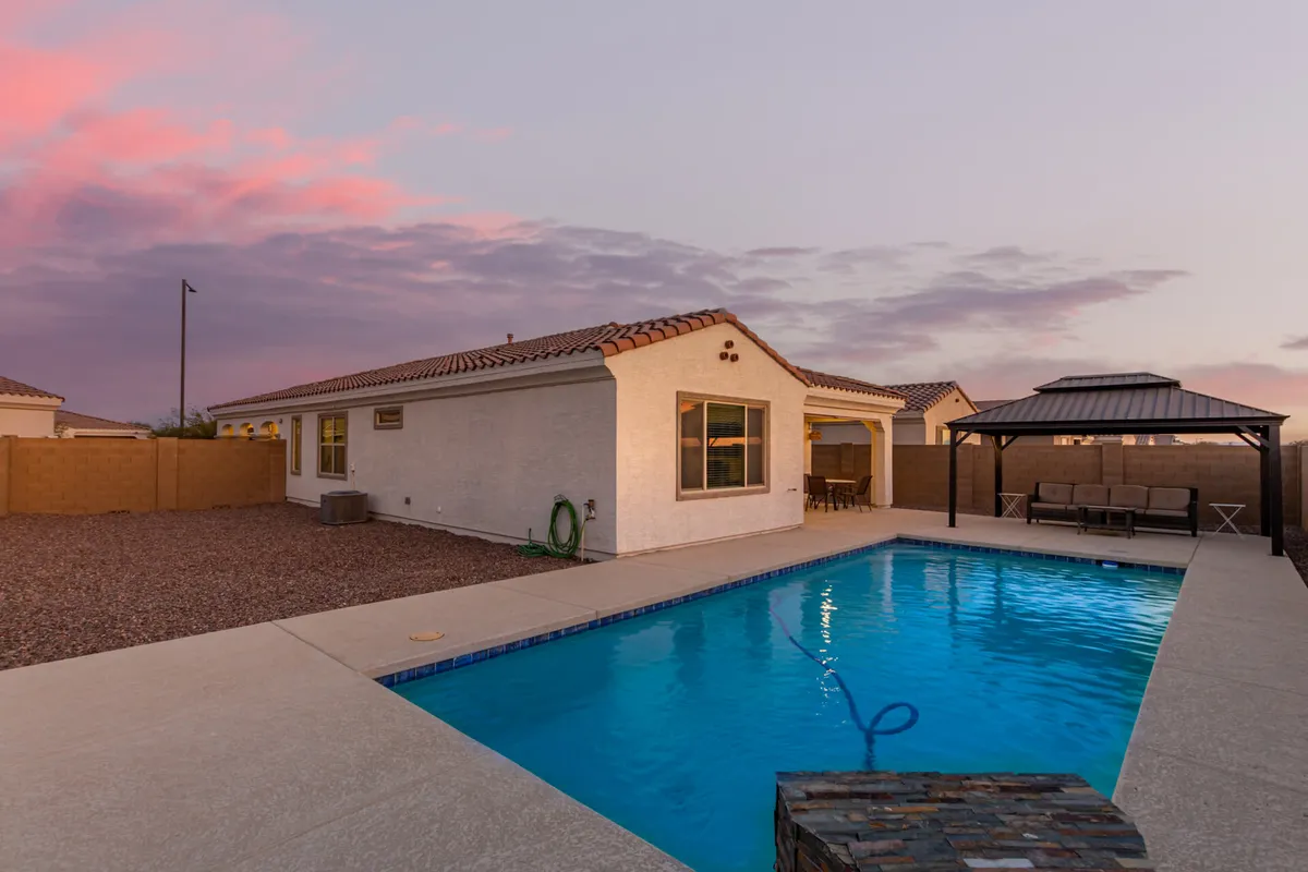 The image showcases the inviting outdoor oasis of a single-family house in Buckeye, Arizona, with a sparkling swimming pool surrounded by lush palm trees, a cozy gazebo perfect for lounging or dining, and the modern Southwestern exterior of the home. The yard is beautifully landscaped, setting the scene for both tranquil mornings and festive evenings by the pool.
