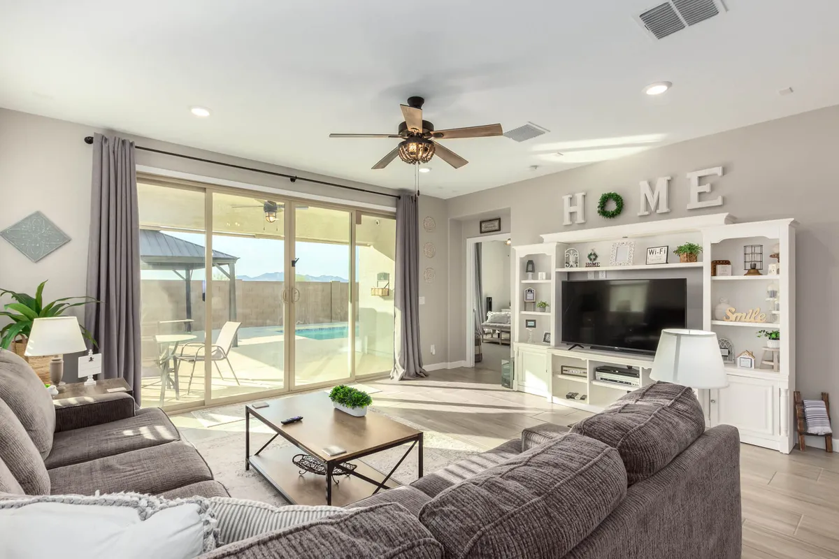 A sunlit living room featuring a comfortable grey sofa facing a large TV unit. Sliding glass doors open to the outdoors, flooding the space with natural light, while a wall displays a decorative 'HOME' sign, adding a touch of warmth. The room is styled with modern Southwestern accents and arranged for group relaxation or lively conversation.