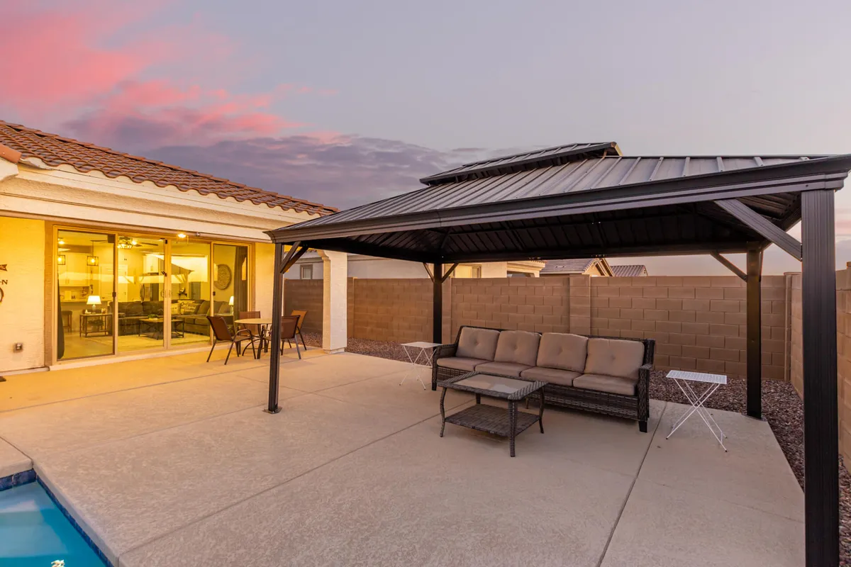 A serene backyard scene featuring a sparkling swimming pool, a shaded gazebo, and a glimpse inside through large glass doors. The area is surrounded by landscaped greenery and palm trees, creating an inviting oasis for relaxation and social gatherings.